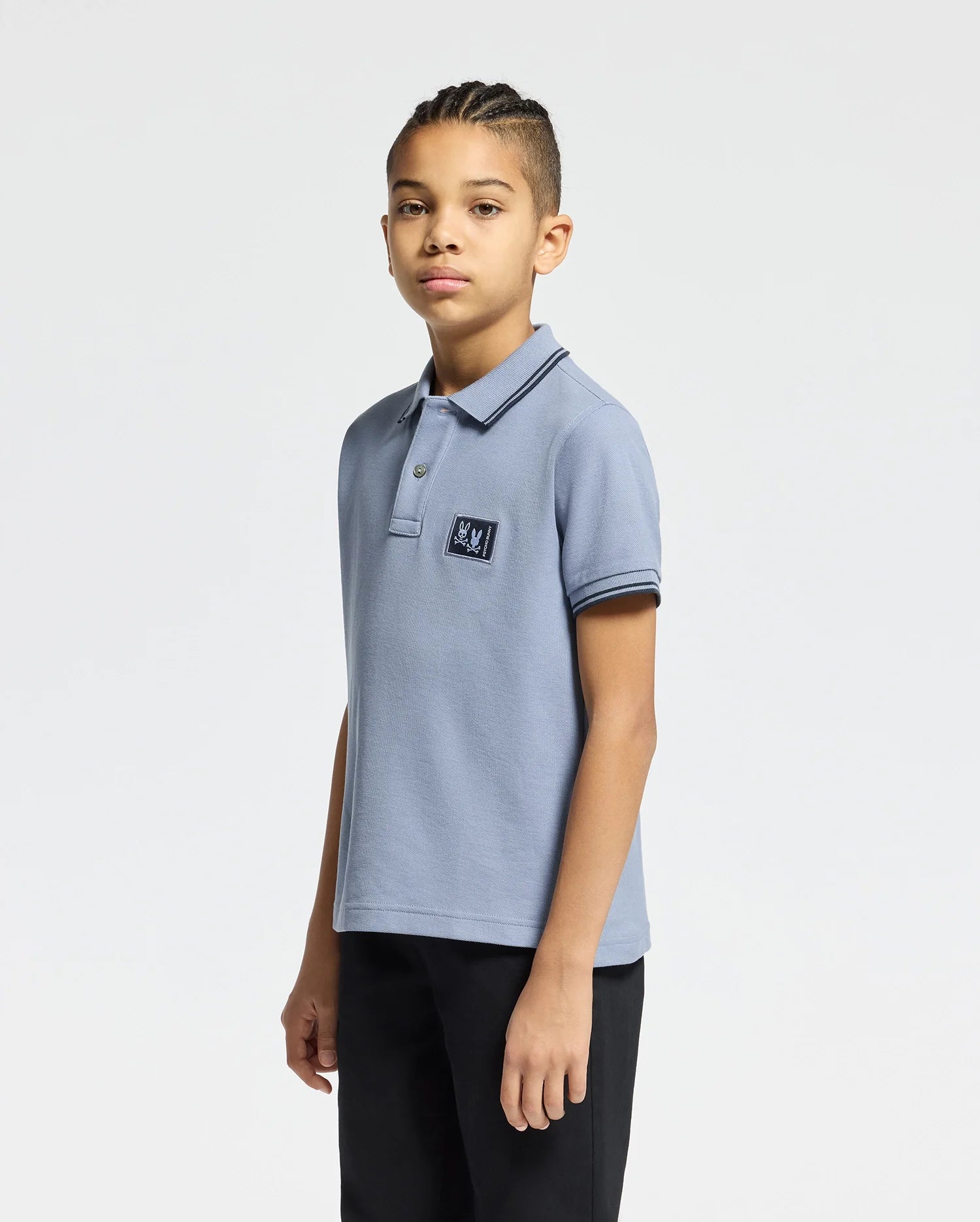 A young person with braided hair wears a Psycho Bunny KIDS MONROE PIQUE POLO in light blue, featuring a chest logo appliqué and regular fit. They're paired with black pants, standing relaxed against a plain white background.
