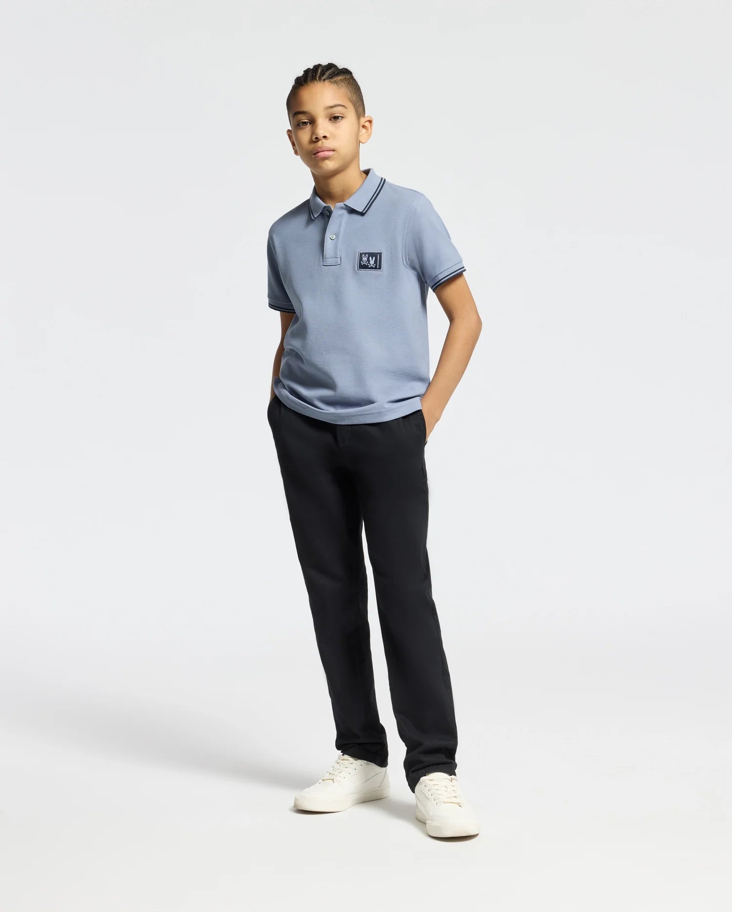 A young person stands confidently in a regular fit light blue Psycho Bunny KIDS MONROE PIQUE POLO (B0K964D200), black pants, and white sneakers. Their hands are in pockets, with a neutral expression against a plain, light background.