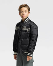 A young boy stands confidently wearing the Psycho Bunny KIDS FRASER FASHION PUFFER - B0N338E200, a trendy black jacket with white and black striped cuffs and collar. The cursive text on the chest adds a touch of style. With light brown hair, he looks directly at the camera against a plain gray background.