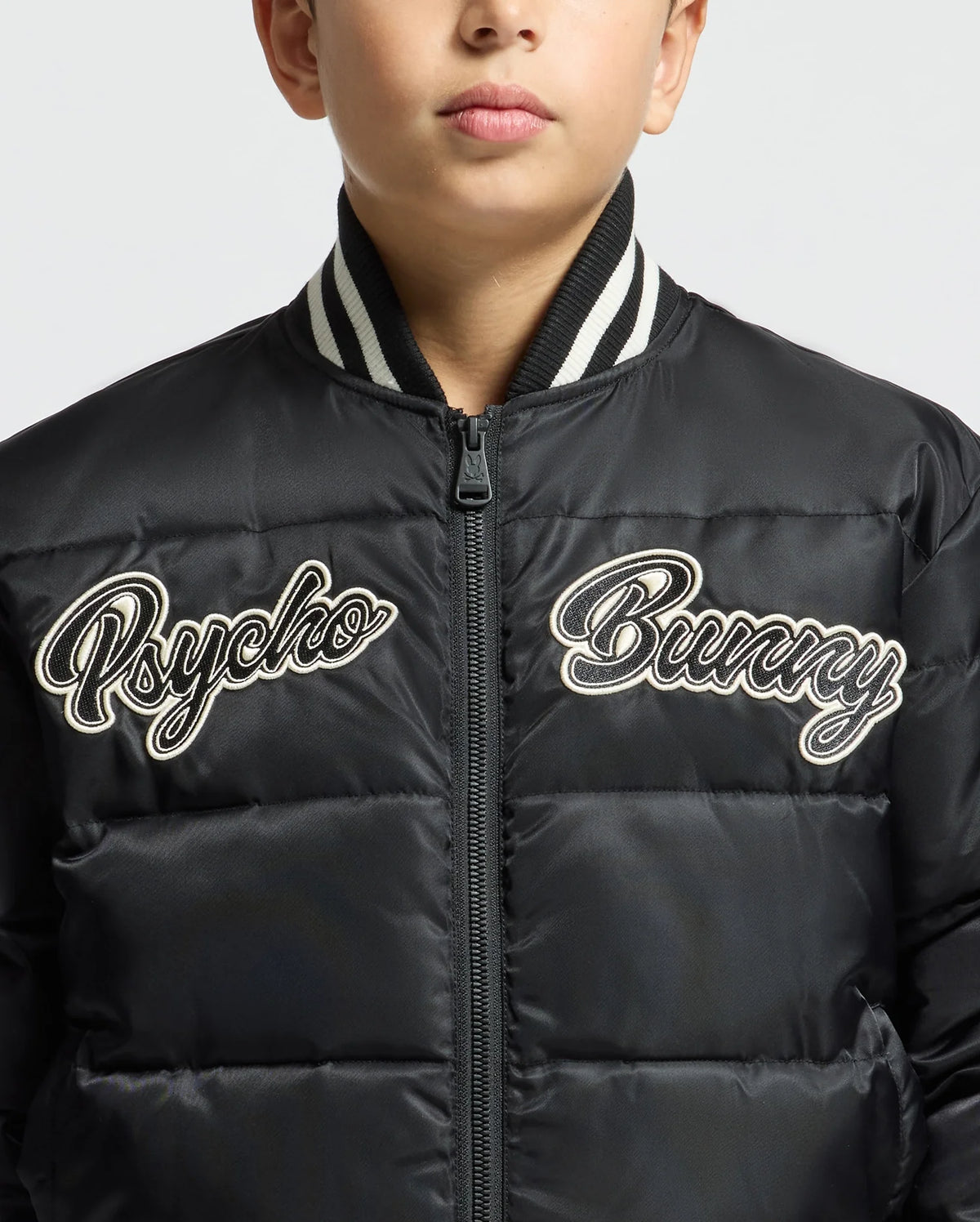 A boy wears the KIDS FRASER FASHION PUFFER - B0N338E200 from Psycho Bunny, a trendy black puffer jacket with white embroidered text "Psycho Bunny" on the front. This warm kids' fashion piece, featuring a ribbed collar, keeps him cozy. His face is partially visible against a plain background.