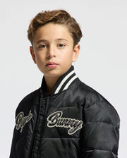 A young boy with short brown hair is wearing a stylish KIDS FRASER FASHION PUFFER - B0N338E200 by Psycho Bunny, featuring "Bunny" and "Royce" embroidery. The puffer jacket boasts a distinctive white and black striped collar. Perfectly capturing the essence of warm kids' fashion, he gazes directly at the camera against a plain white backdrop.
