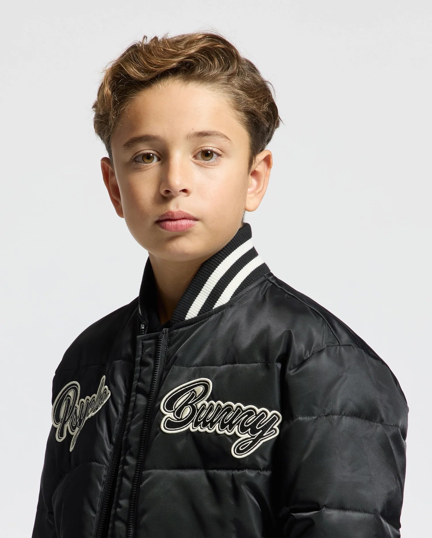 A young boy with short brown hair is wearing a stylish KIDS FRASER FASHION PUFFER - B0N338E200 by Psycho Bunny, featuring "Bunny" and "Royce" embroidery. The puffer jacket boasts a distinctive white and black striped collar. Perfectly capturing the essence of warm kids' fashion, he gazes directly at the camera against a plain white backdrop.