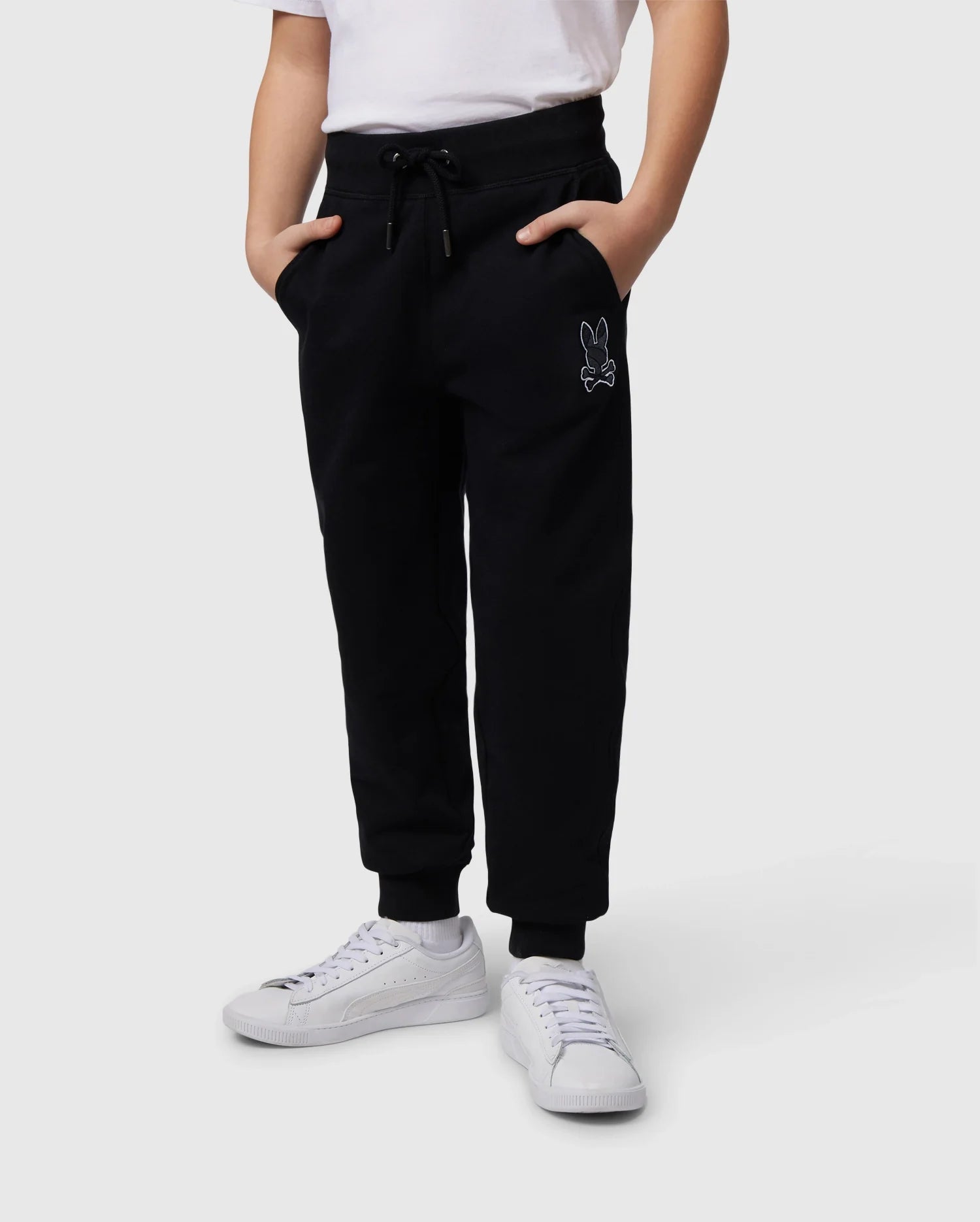 A person wearing the Psycho Bunny KIDS LENOX REGULAR FIT SWEATPANT, featuring black cotton fleece material and an embroidered bunny logo on the upper left thigh, with hands in the pockets. They are paired with a white t-shirt and white sneakers, standing against a plain gray background.