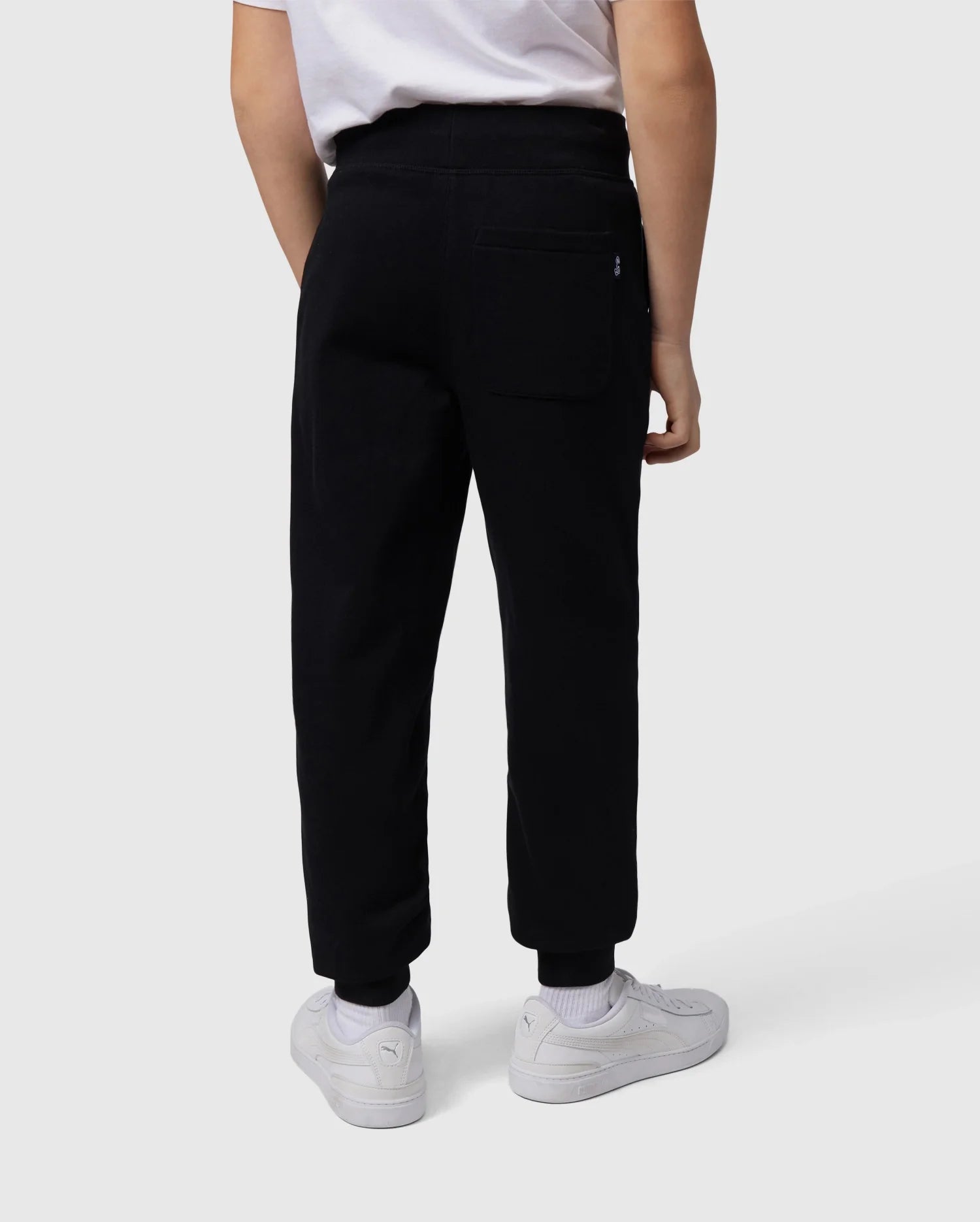 A back view of a person wearing a white T-shirt, white sneakers, and Psycho Bunny's KIDS LENOX REGULAR FIT SWEATPANT in black made from soft cotton fleece with an embroidered logo on the back pocket. The person's right arm is resting by their side against a plain white background.