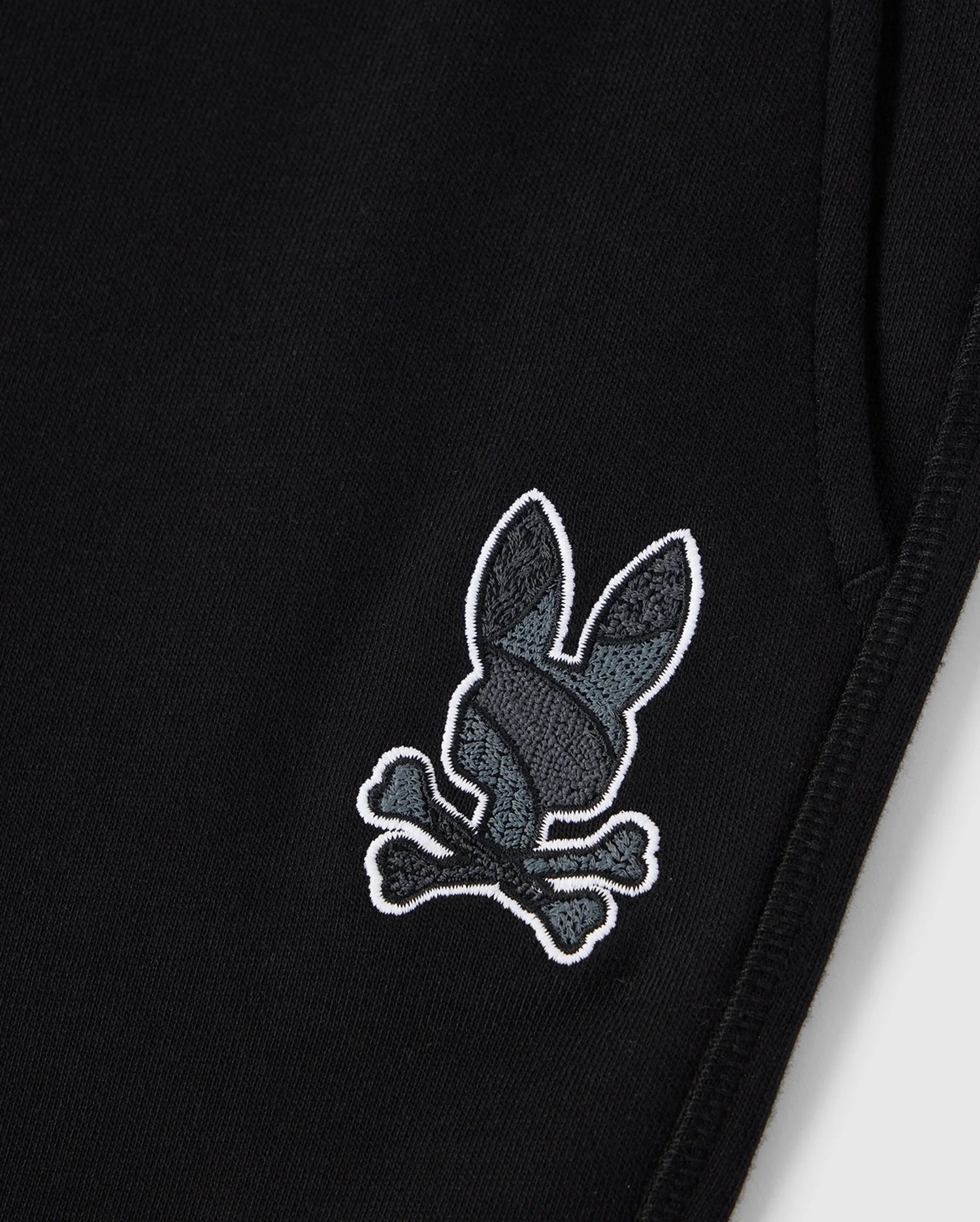 A close-up view of the KIDS LENOX REGULAR FIT SWEATPANT by Psycho Bunny, in black cotton fleece. The sweatpants feature an embroidered logo of a gray rabbit head with black detailing and crossed bones underneath, mimicking the traditional pirate skull and crossbones symbol.