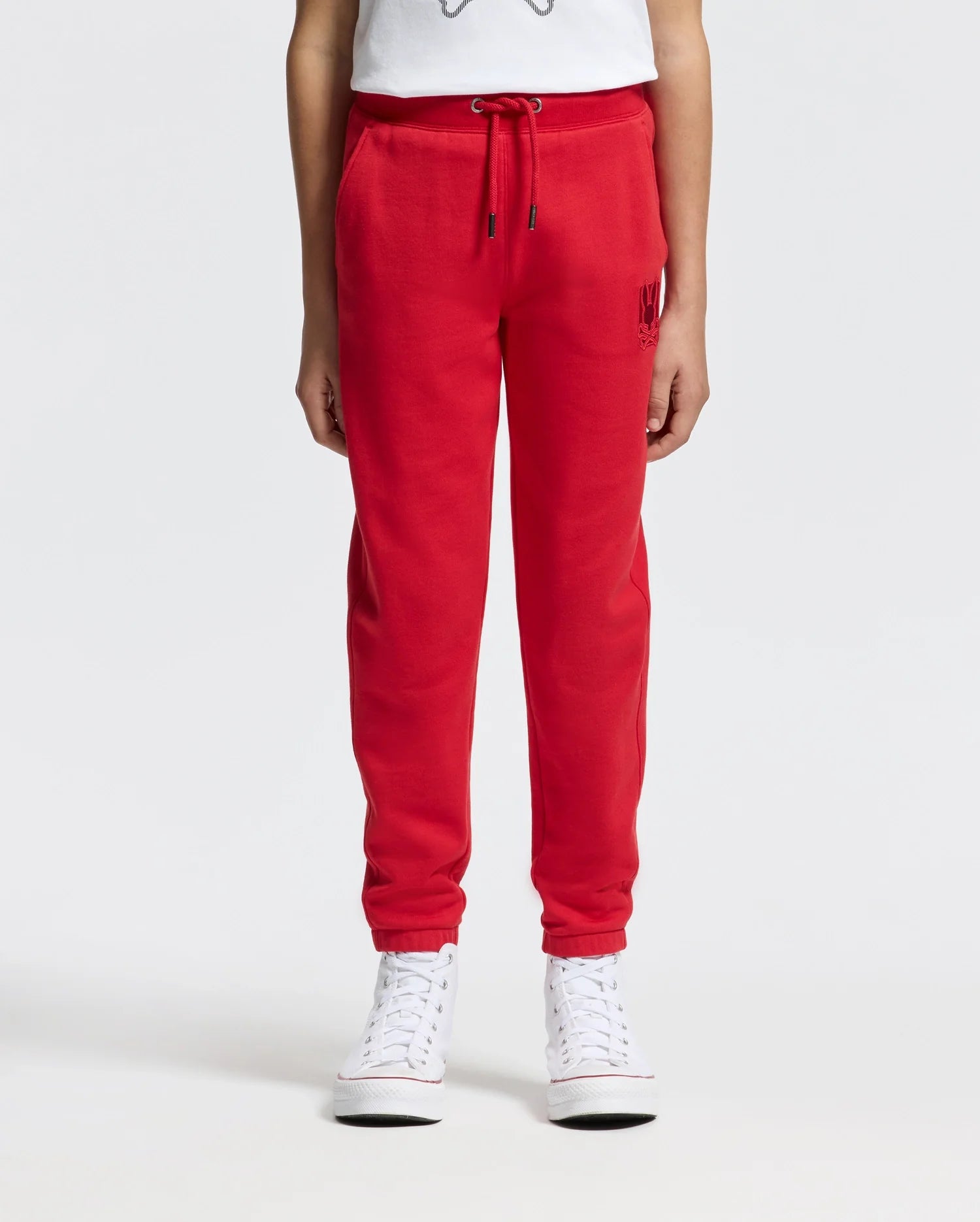 A person wearing vibrant red KIDS PIERCE ESSENTIAL FRENCH TERRY SWEATPANTS by Psycho Bunny, paired with white high-top sneakers, stands against a plain white background. These premium kids' joggers feature an elastic waistband, a drawstring, and side pockets.