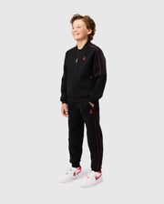 A young boy stands smiling, dressed in the KIDS TRAVIS TRACK PANT - B0P143D200 from Psycho Bunny. He has his hands in the elastic and comfortable pants pockets of his black zip-up tracksuit with red accents. The outfit features a small red logo on the chest and thigh. He complements his look with white sneakers featuring red details, all against a plain grey background.