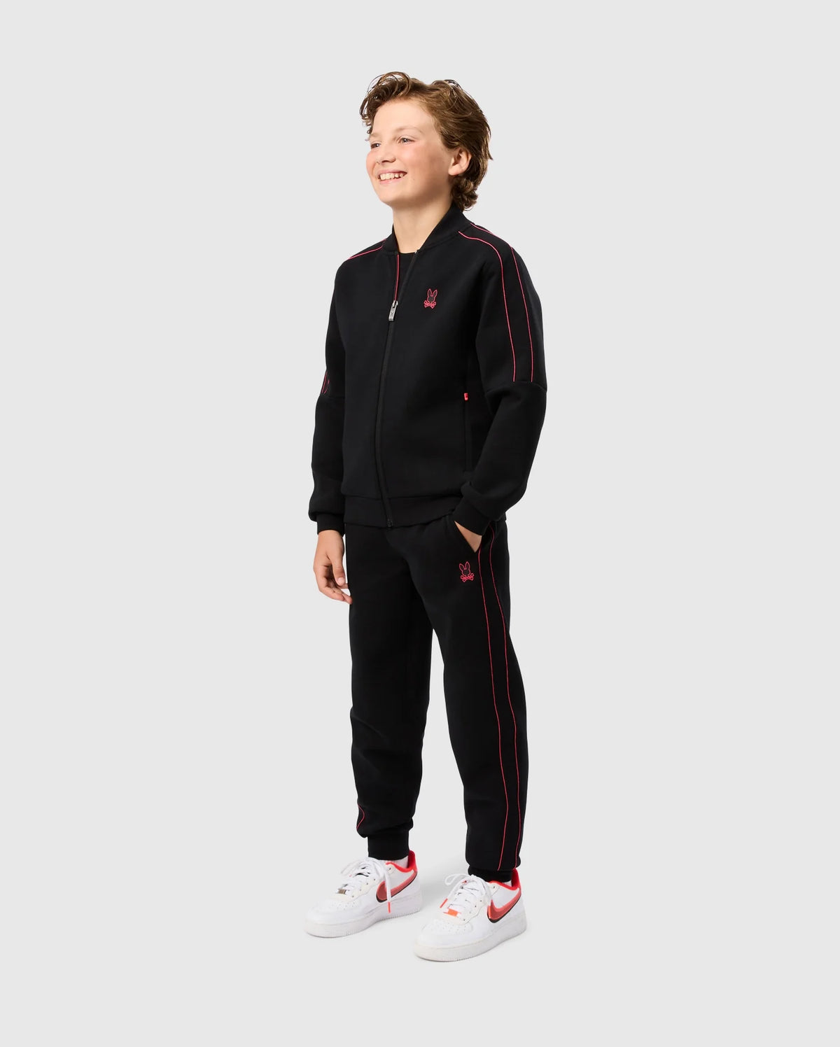 A young boy stands smiling, dressed in the KIDS TRAVIS TRACK PANT - B0P143D200 from Psycho Bunny. He has his hands in the elastic and comfortable pants pockets of his black zip-up tracksuit with red accents. The outfit features a small red logo on the chest and thigh. He complements his look with white sneakers featuring red details, all against a plain grey background.
