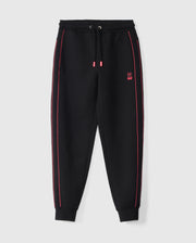 The Psycho Bunny KIDS TRAVIS TRACK PANT - B0P143D200 features black elastic and comfortable kids' track pants with a drawstring waist and red accents, including contrast chainstitch stripes along the sides and red-tipped drawstrings. A small red bunny logo is embroidered on the left thigh. The cuffs and waistband are elasticated, presented against a white background.