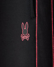 Close-up of the black fabric of the KIDS TRAVIS TRACK PANT - B0P143D200 by Psycho Bunny, featuring a pink bunny skull and crossbones logo. The elastic and comfortable fabric showcases contrast chainstitch stripes running vertically to the right of the logo.