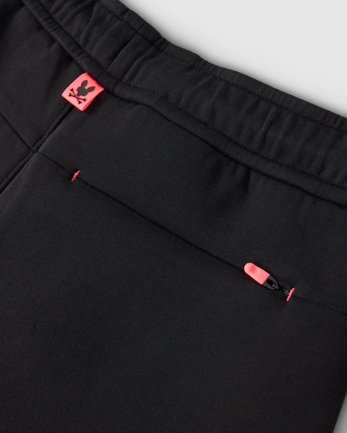Close-up of the KIDS TRAVIS TRACK PANT - B0P143D200 by Psycho Bunny, showcasing an elastic and comfortable waistband. These black pants include a back pocket with a pink zipper pull and a small pink tag with the Psycho Bunny logo near the waistband. The fabric is smooth and well-stitched, accented by contrast chainstitch stripes for added style.