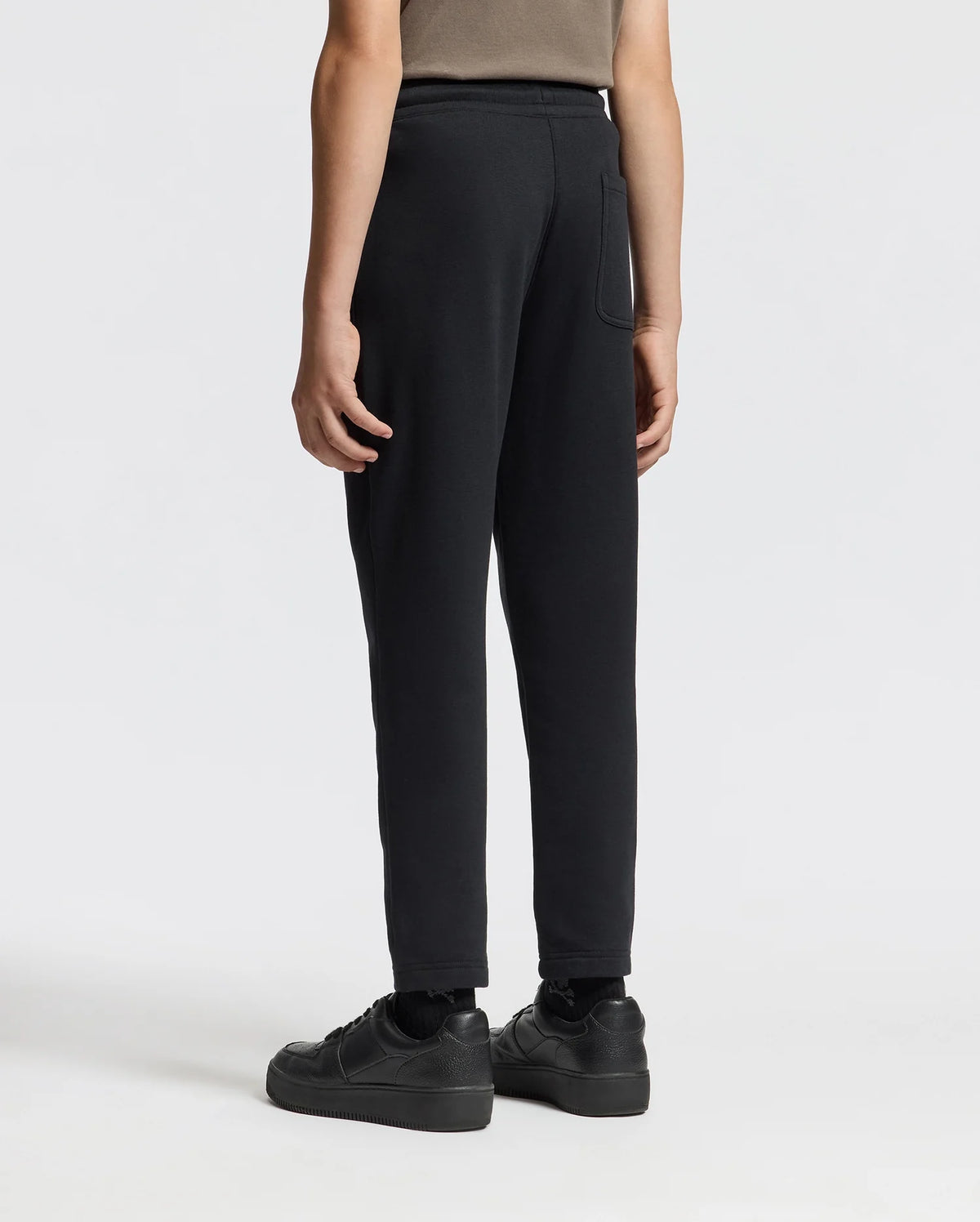 A person is standing against a plain white background, viewed from the back, wearing a brown shirt, KIDS ALEXANDER SWEATPANT - B0P155E200 from Psycho Bunny in black, and black sneakers.