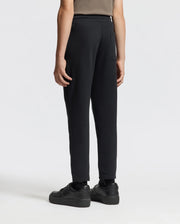 A person is standing against a plain white background, viewed from the back, wearing a brown shirt, KIDS ALEXANDER SWEATPANT - B0P155E200 from Psycho Bunny in black, and black sneakers.