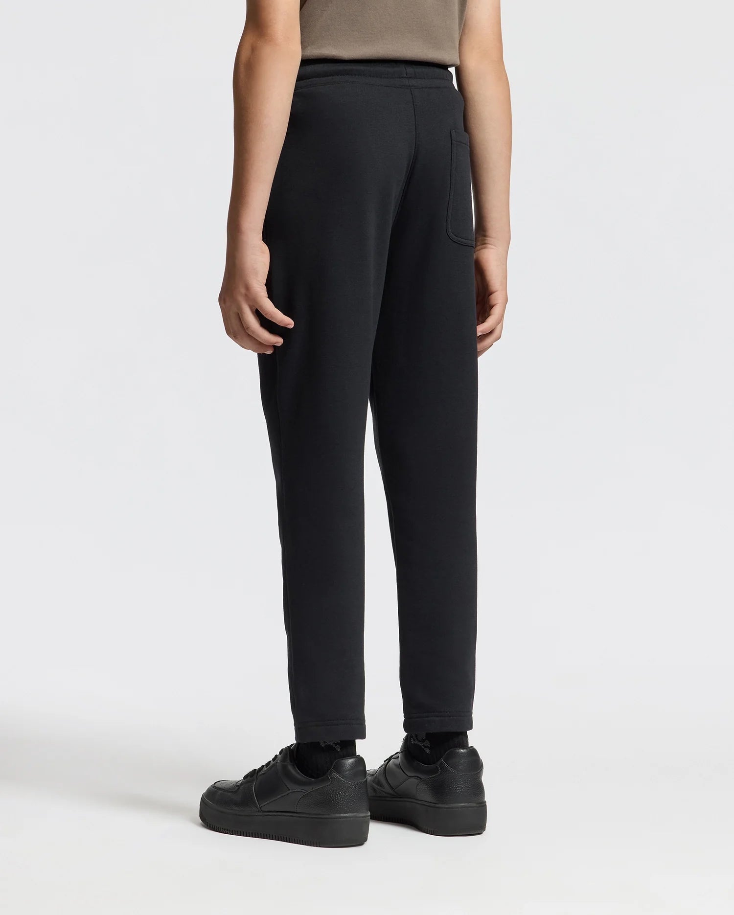 A person is standing against a plain white background, viewed from the back, wearing a brown shirt, KIDS ALEXANDER SWEATPANT - B0P155E200 from Psycho Bunny in black, and black sneakers.