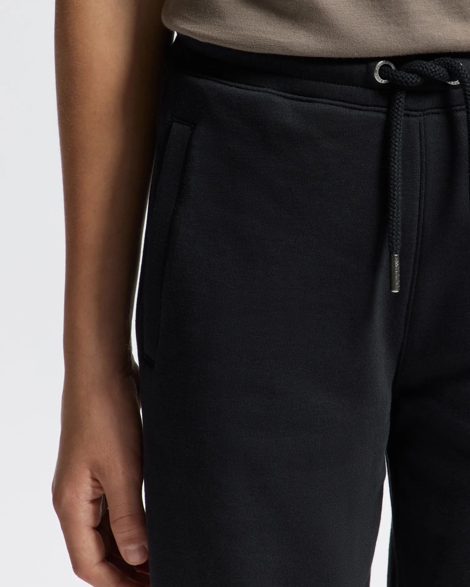 A person is wearing the Psycho Bunny KIDS ALEXANDER SWEATPANT - B0P155E200 in black, featuring a side pocket. Their right hand is partially visible, resting by their side. The sweatpants are paired with a beige top, all set against a plain white background.