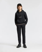 A young person stands confidently, dressed in a black hoodie featuring a rabbit skull design, kids' black sweatpants from the Psycho Bunny collection, a matching cap, and sneakers. The sleek cotton blend of the KIDS ALEXANDER SWEATPANT - B0P155E200 enhances their edgy style against the plain white background.