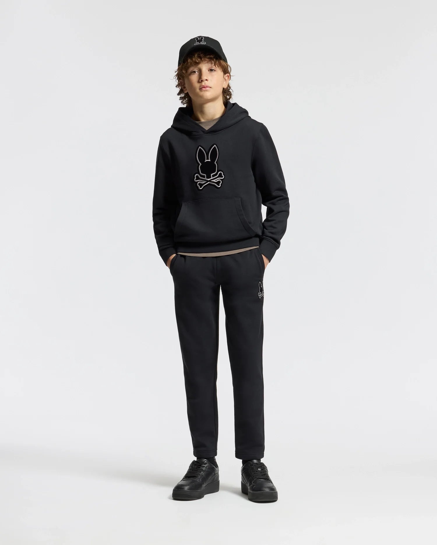 A young person stands confidently, dressed in a black hoodie featuring a rabbit skull design, kids' black sweatpants from the Psycho Bunny collection, a matching cap, and sneakers. The sleek cotton blend of the KIDS ALEXANDER SWEATPANT - B0P155E200 enhances their edgy style against the plain white background.
