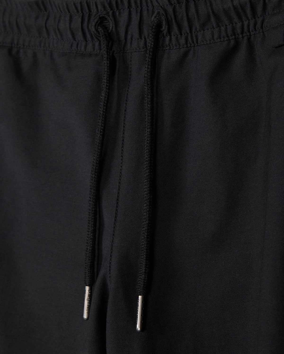 A close-up image of the black KIDS MADISON COMMUTER PANT - B0P395Z1WB by Psycho Bunny, focusing on the waistband. The pants feature an elastic waistband with drawstrings that have metal tips. The fabric appears to be smooth and solid in color.