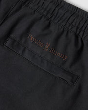 Close-up of a black KIDS MADISON COMMUTER PANT - B0P395Z1WB from Psycho Bunny, featuring an elastic waistband and a pocket with subtle brown embroidery. The embroidery includes the text "Psycho Bunny" with a small bunny logo positioned between the two words. Zippered expansion vents add functionality to this stylish piece.