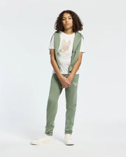 A person with curly hair wears a light green hoodie, a white T-shirt with a rabbit design, and matching KIDS DESERT EMBROIDERED SWEATPANTS by Psycho Bunny. They finish the look with white sneakers against a plain white background.