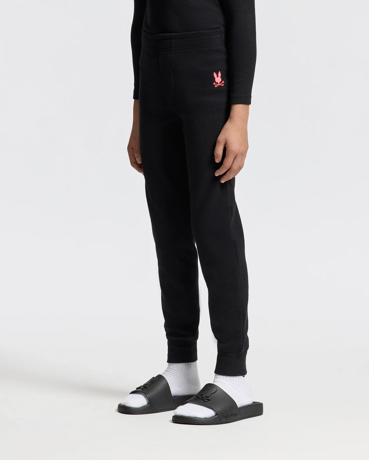 A person is standing against a plain background wearing the cozy KIDS VAL WAFFLE PANT by Psycho Bunny, in black, featuring a pink logo on the upper thigh. They are also wearing black slide sandals paired with white socks.