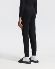 A person wearing black KIDS VAL WAFFLE PANT - B0P673E200 by Psycho Bunny, paired with a matching black long-sleeve top, white socks, and black slides stands facing away against a plain white background. The focus is on the clothing.