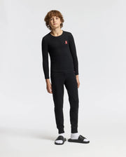Someone with curly hair is wearing the Psycho Bunny KIDS VAL WAFFLE PANT in a cozy style, along with a black long-sleeve top featuring a pink design on the pocket. They're also sporting black slide sandals paired with white socks, all set against a plain white background.