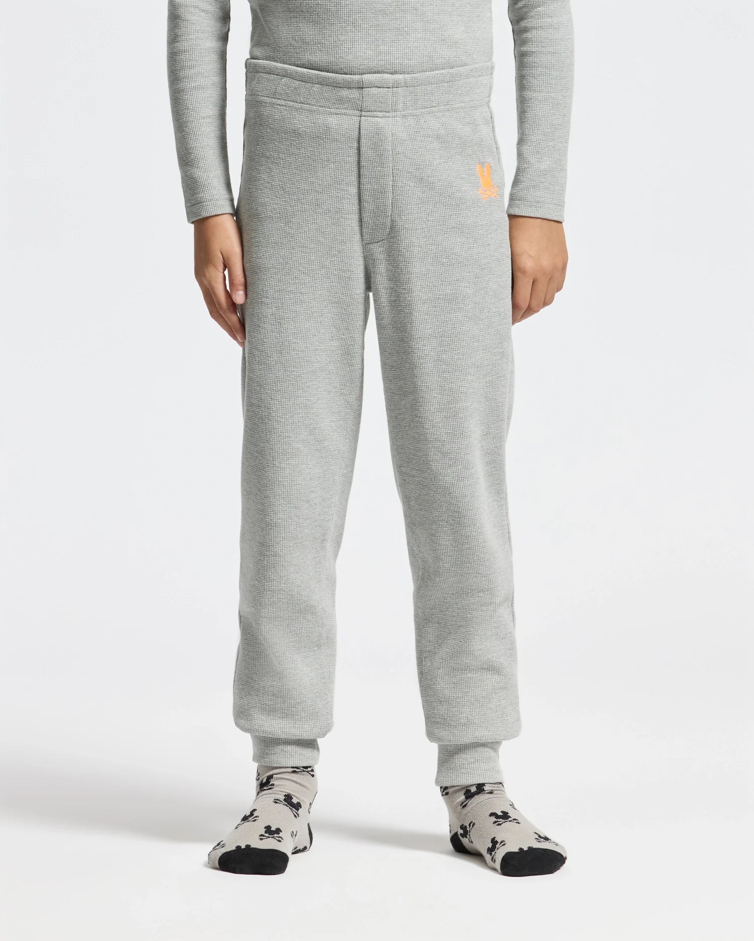A person is comfortably standing, wearing the gray KIDS VAL WAFFLE PANT - B0P673E200 by Psycho Bunny, featuring a small orange logo near the pocket. They also have on a gray long-sleeve shirt and gray socks with a black pattern against a plain white background.