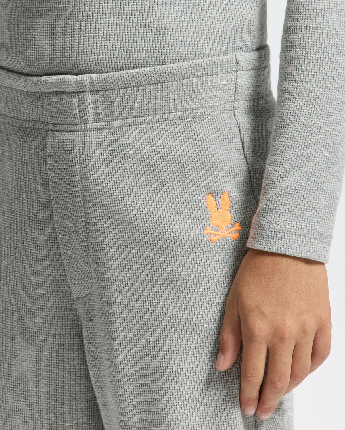 A close-up view features a person in the KIDS VAL WAFFLE PANT by Psycho Bunny, showcasing a cozy textured lounge set with long sleeves. The Pima cotton pants are gray, highlighted by an embroidered small orange bunny logo on the side, while the person's hand rests casually at their side.