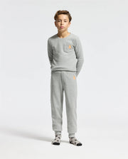 A young boy stands against a plain white background wearing the Psycho Bunny KIDS VAL WAFFLE PANT - B0P673E200, a light gray shirt and cozy waffle pants set. The outfit includes a small orange emblem on the chest and hip for a pop of color. He completes his look with patterned socks, epitomizing casual yet stylish comfort.