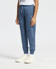 A person wearing a white T-shirt, comfortable Kids Levy Essential Fleece Sweatpants by Psycho Bunny featuring a small embroidered logo near the drawstring kangaroo pocket, white socks, and white sneakers stands against a plain background. Only the lower half of the body is visible.