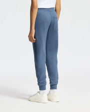 A person is standing sideways, facing away while wearing a white shirt, a pair of Psycho Bunny KIDS LEVY ESSENTIAL FLEECE SWEATPANT - B0P721D200 in blue with a drawstring kangaroo pocket, and white sneakers. The background is a plain light color. This ensemble epitomizes comfortable casual wear.