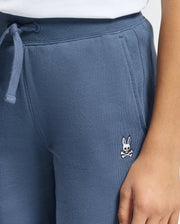 A person is dressed in the KIDS LEVY ESSENTIAL FLEECE SWEATPANT by Psycho Bunny, featuring a comfortable cotton fleece fabric and a waistband drawstring. The pants display an embroidered rabbit head with crossbones logo. The person's hand rests along the side of the sweatpants, highlighting their casual style.