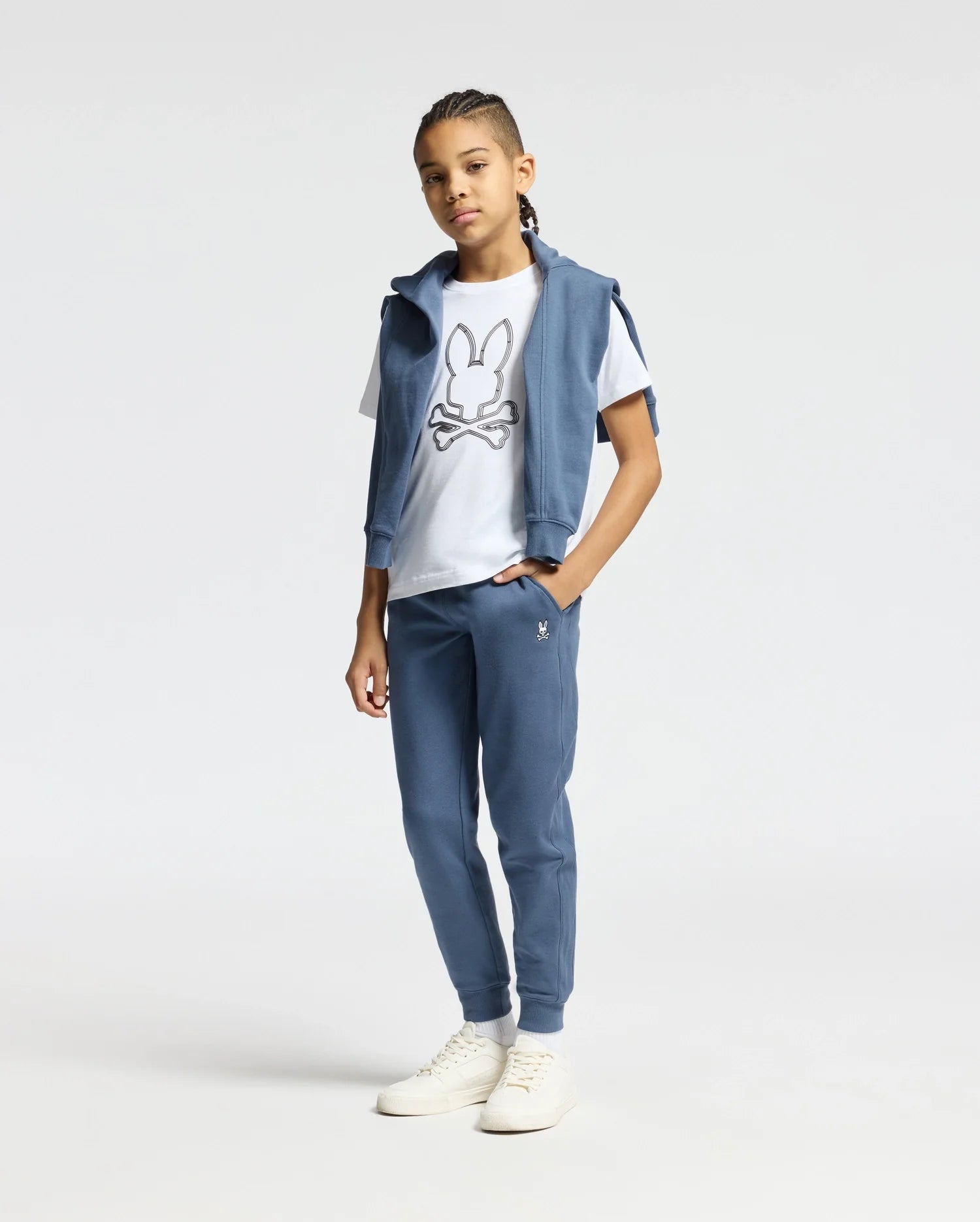 A young person with braids stands confidently, wearing a white T-shirt featuring a cartoon design and a blue vest casually draped over their shoulders. They are also sporting the KIDS LEVY ESSENTIAL FLEECE SWEATPANT by Psycho Bunny for maximum comfort. The outfit is completed with white sneakers, set against a plain, light-colored background.