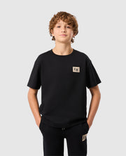 A young person with curly hair stands against a light gray background, wearing a black T-shirt with a small beige square emblem and Psycho Bunny's Kids Monroe Twill Patch Fleece Sweatpant. Their hands are in their pockets, exuding casual confidence as they gaze straight ahead.