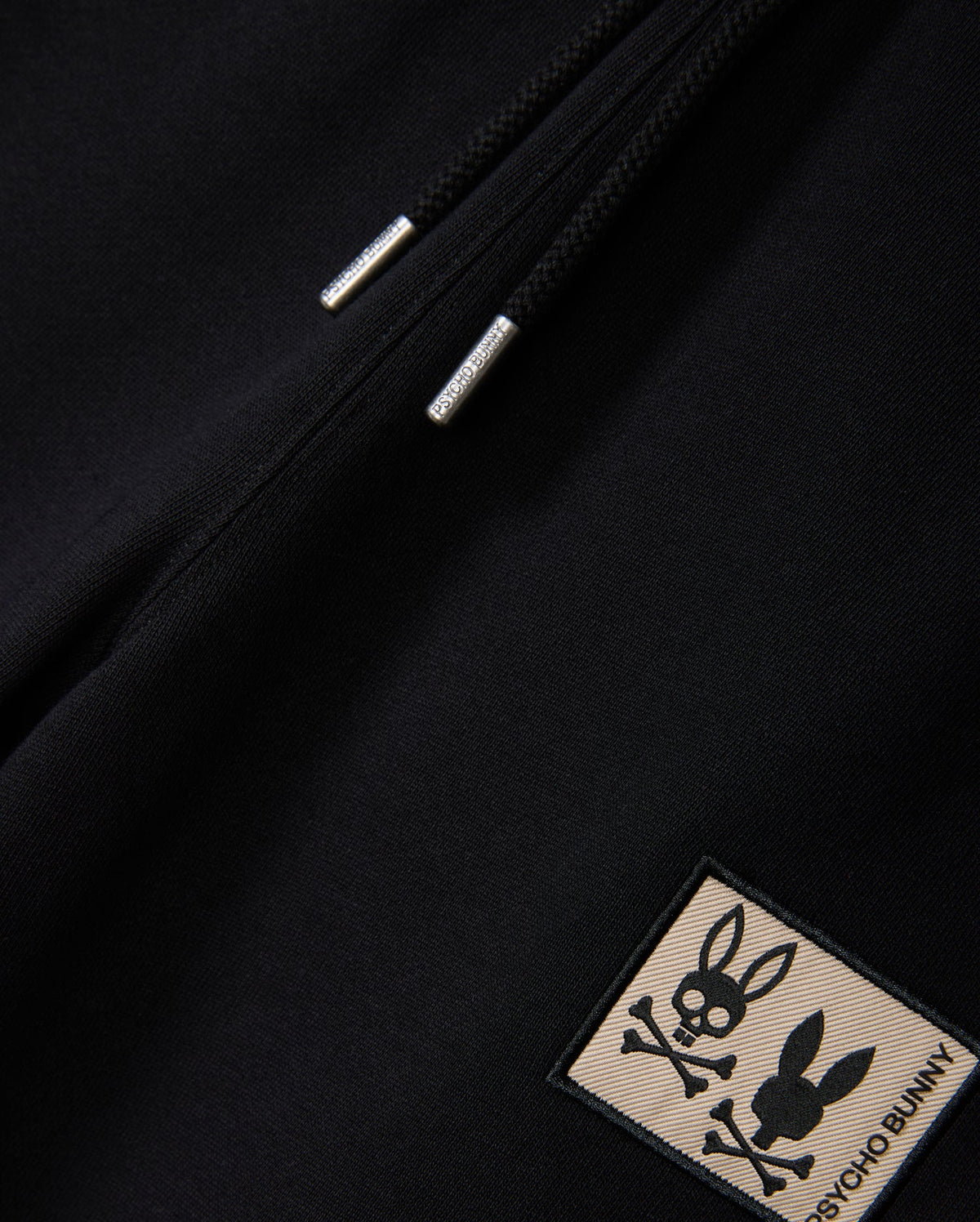 A close-up of the KIDS MONROE TWILL PATCH FLEECE SWEATPANT in black by Psycho Bunny features a zipper with metal pull tabs and luxury finishes, plus a bunny patch showcasing a stylized bunny skull with crossed bones and the text "Psycho Bunny.