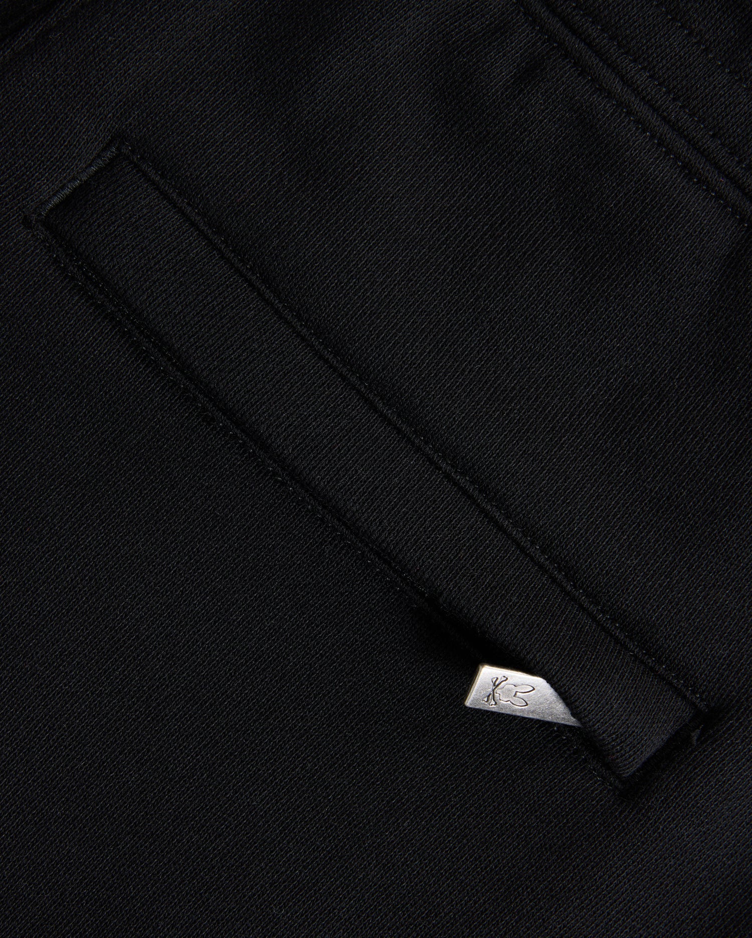A close-up of black Psycho Bunny KIDS MONROE TWILL PATCH FLEECE SWEATPANTS showcases a welt pocket with a white tag featuring the logo. The slightly textured fabric and clean stitching evoke premium craftsmanship.