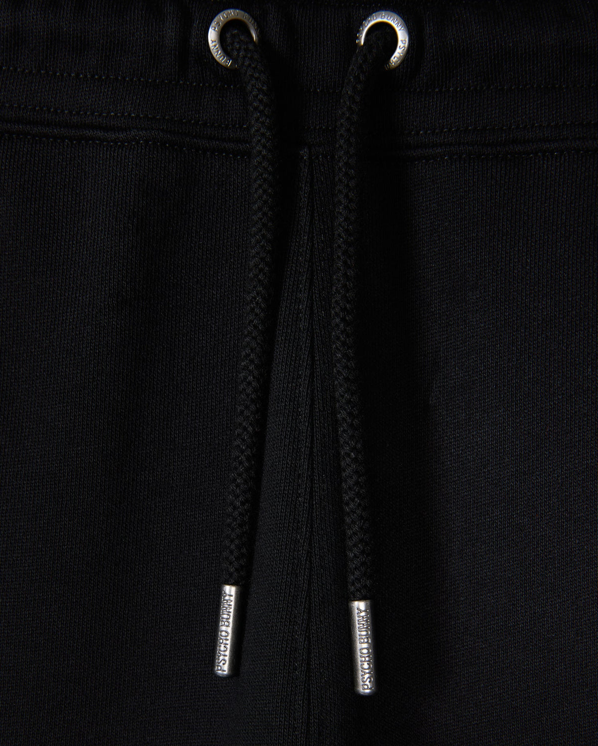 Close-up of black Psycho Bunny KIDS MONROE TWILL PATCH FLEECE SWEATPANT with luxury finishes, including drawstrings with metal tips threaded through metal eyelets at the waistband against a black fabric backdrop.