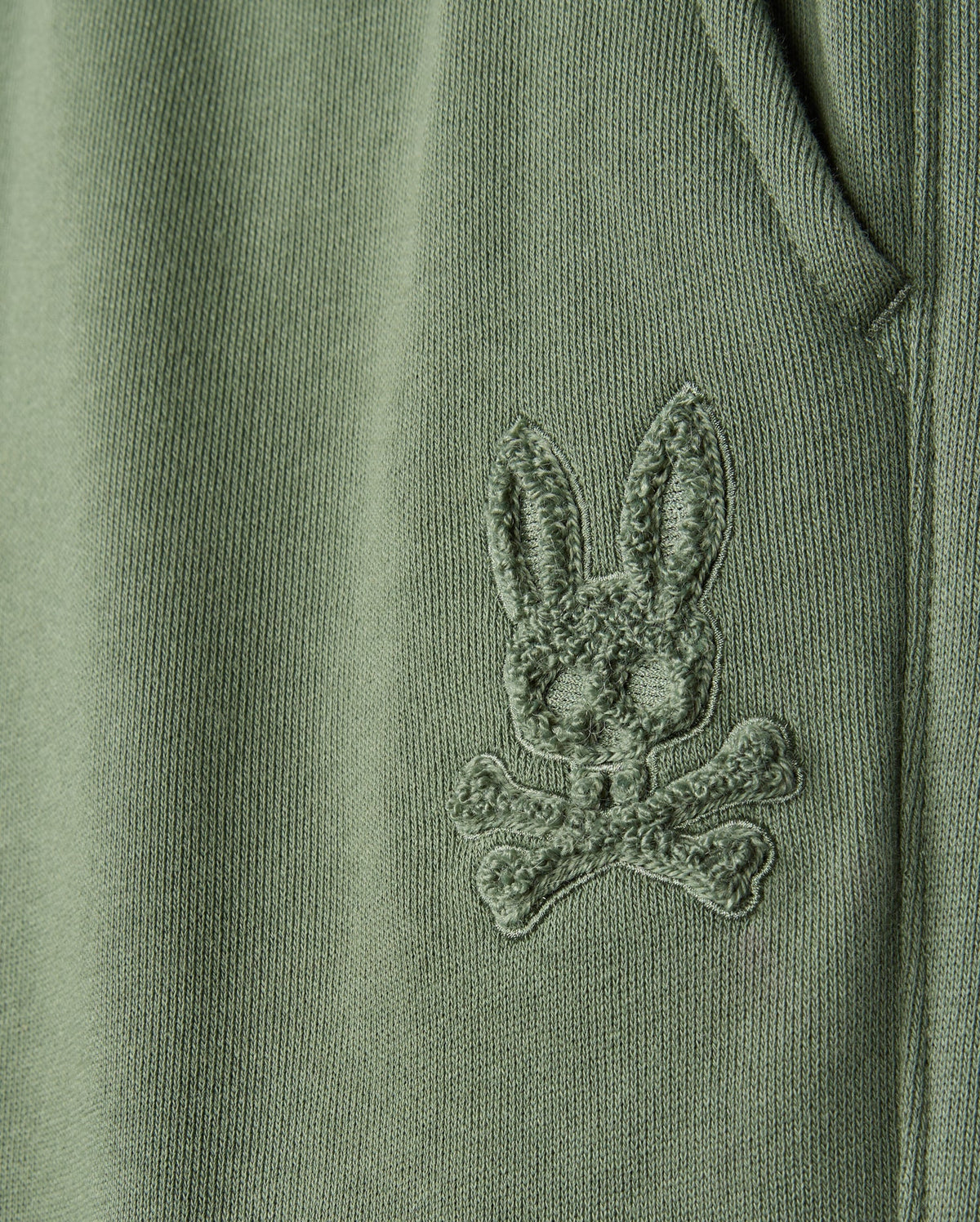 Close-up of a green fabric from the KIDS PACIFIC CHENILLE JOGGER - B0P972D200 by Psycho Bunny, featuring an embroidered bunny's head with crossbones beneath it, reminiscent of a chenille logo patch. The detailed stitching adds a textured appearance to the material.