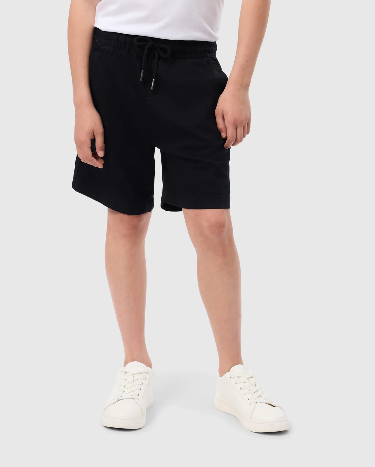 A person is standing against a plain backdrop, seen from the waist down. They are wearing a pair of black KIDS WILLIS STRETCH TENCEL SHORT - B0R239Y1WB by Psycho Bunny, a white T-shirt adorned with an embroidered Bunny logo, and matching white sneakers. Their hands are relaxed at their sides, and the background is light gray.