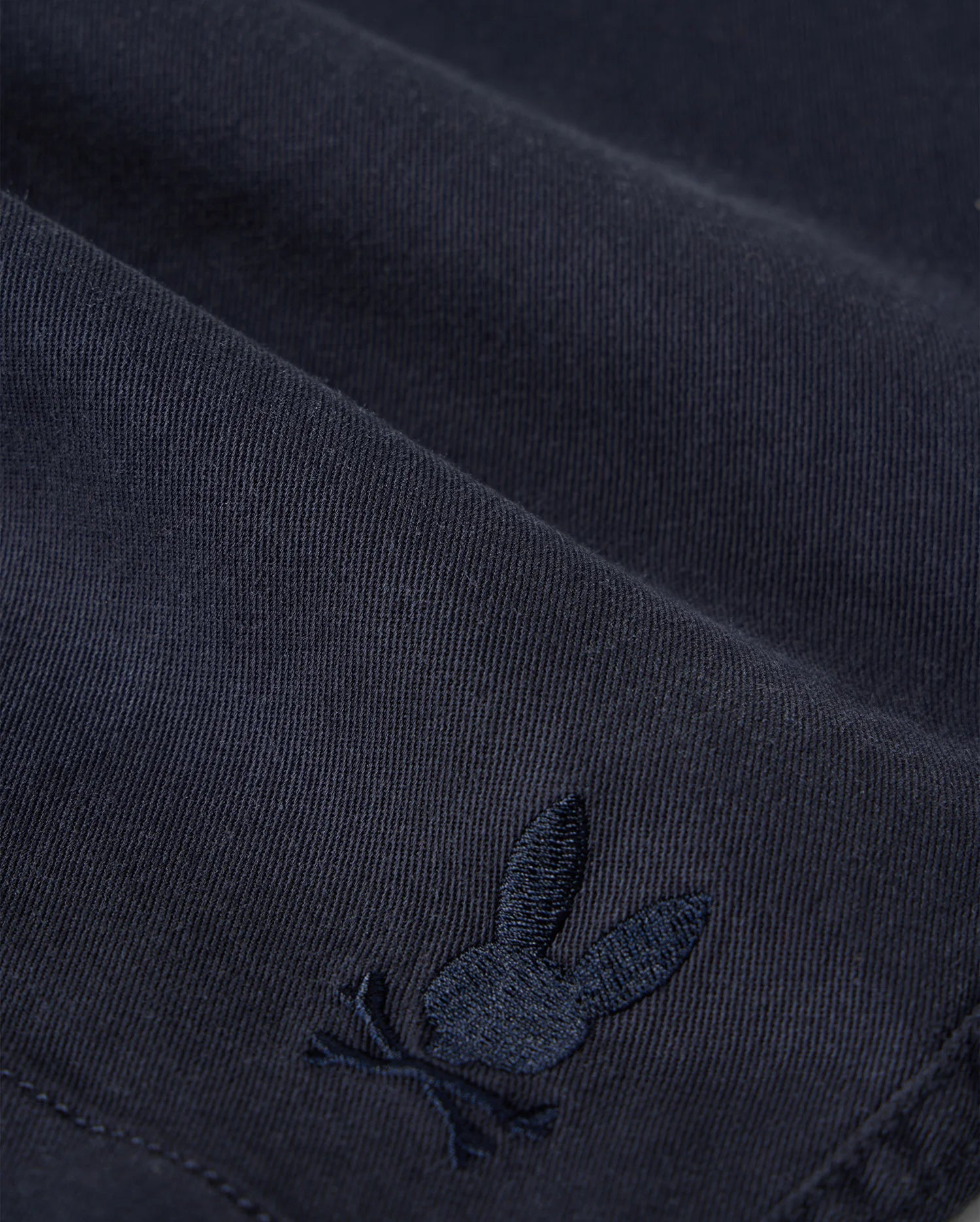 Close-up of Psycho Bunny kids' shorts made from dark KIDS WILLIS STRETCH TENCEL SHORT - B0R239Y1WB twill with a subtle, embroidered logo of a rabbit. The embroidery detail is in a slightly lighter shade of the same color as the fabric.