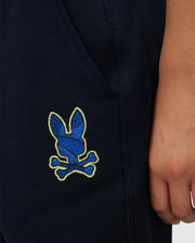 Close-up of a person in Psycho Bunny's KIDS LENOX SWEATSHORT, B0R377B200, wearing dark pants with a blue bunny and crossbones on the side. The naturally breathable fleece ensures comfort for an active lifestyle as their hand rests near the pocket.