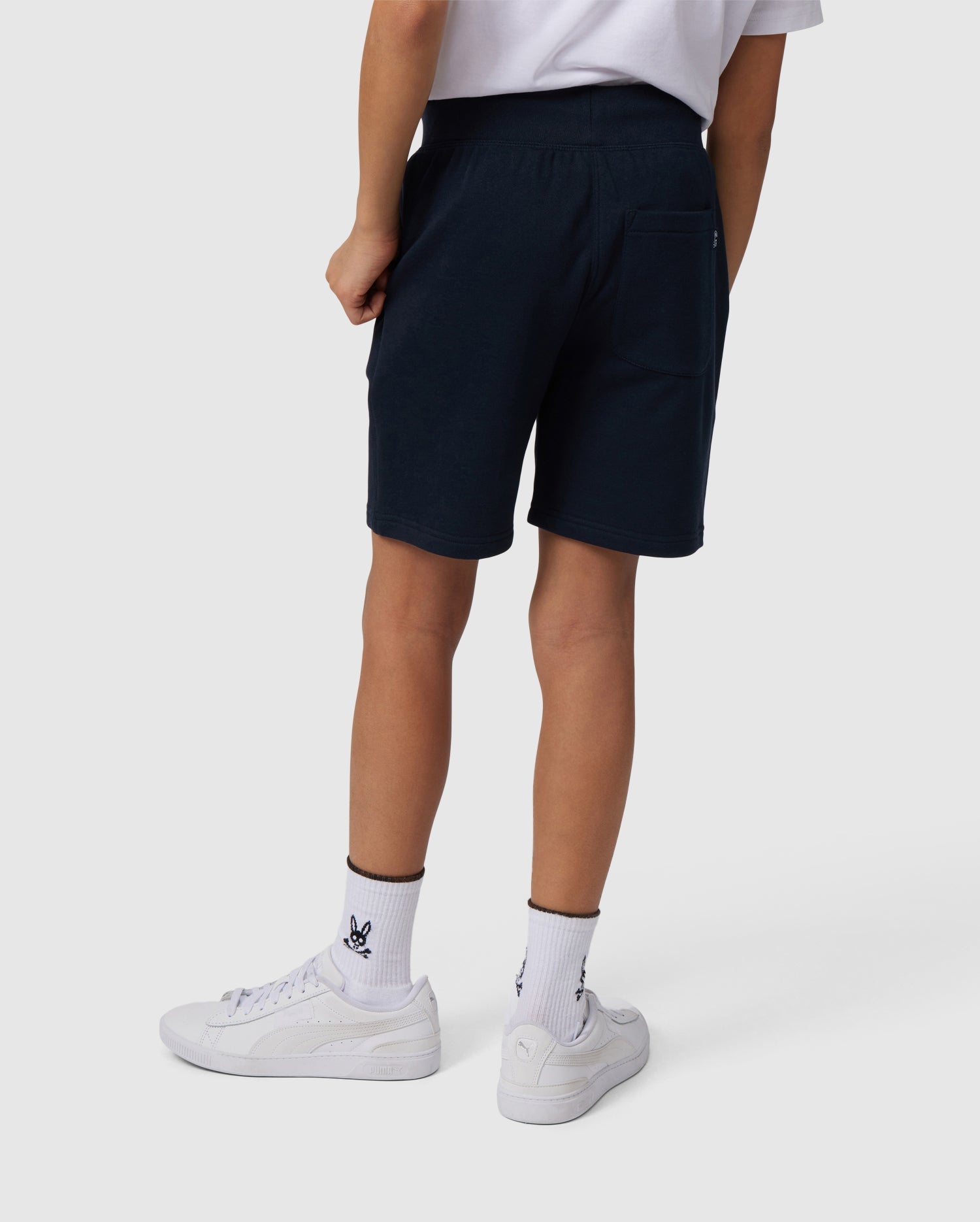 The person, embodying an active lifestyle, wears a white t-shirt and black shorts similar to Psycho Bunny's KIDS LENOX SWEATSHORT - B0R377B200. They complete the look with rabbit-logo white socks and white sneakers, standing against a plain backdrop, ready for movement and adventure.