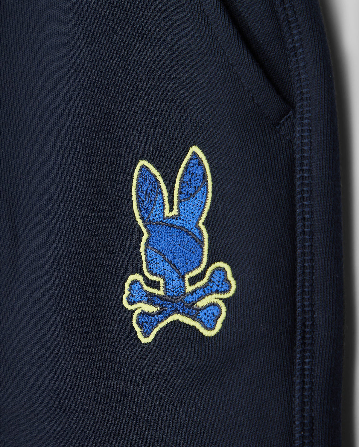 Close-up of Psycho Bunny's navy kids' Lenox sweatshort (B0R377B200) showing a blue bunny head patch with yellow outline and crossed bones near the pocket seam. Made from breathable fleece, it's ideal for an active lifestyle.