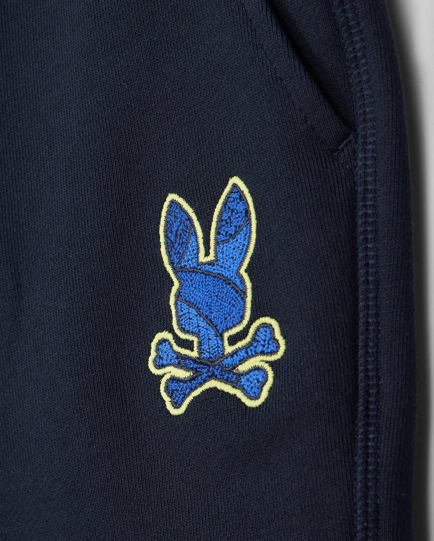 Close-up of Psycho Bunny's navy kids' Lenox sweatshort (B0R377B200) showing a blue bunny head patch with yellow outline and crossed bones near the pocket seam. Made from breathable fleece, it's ideal for an active lifestyle.