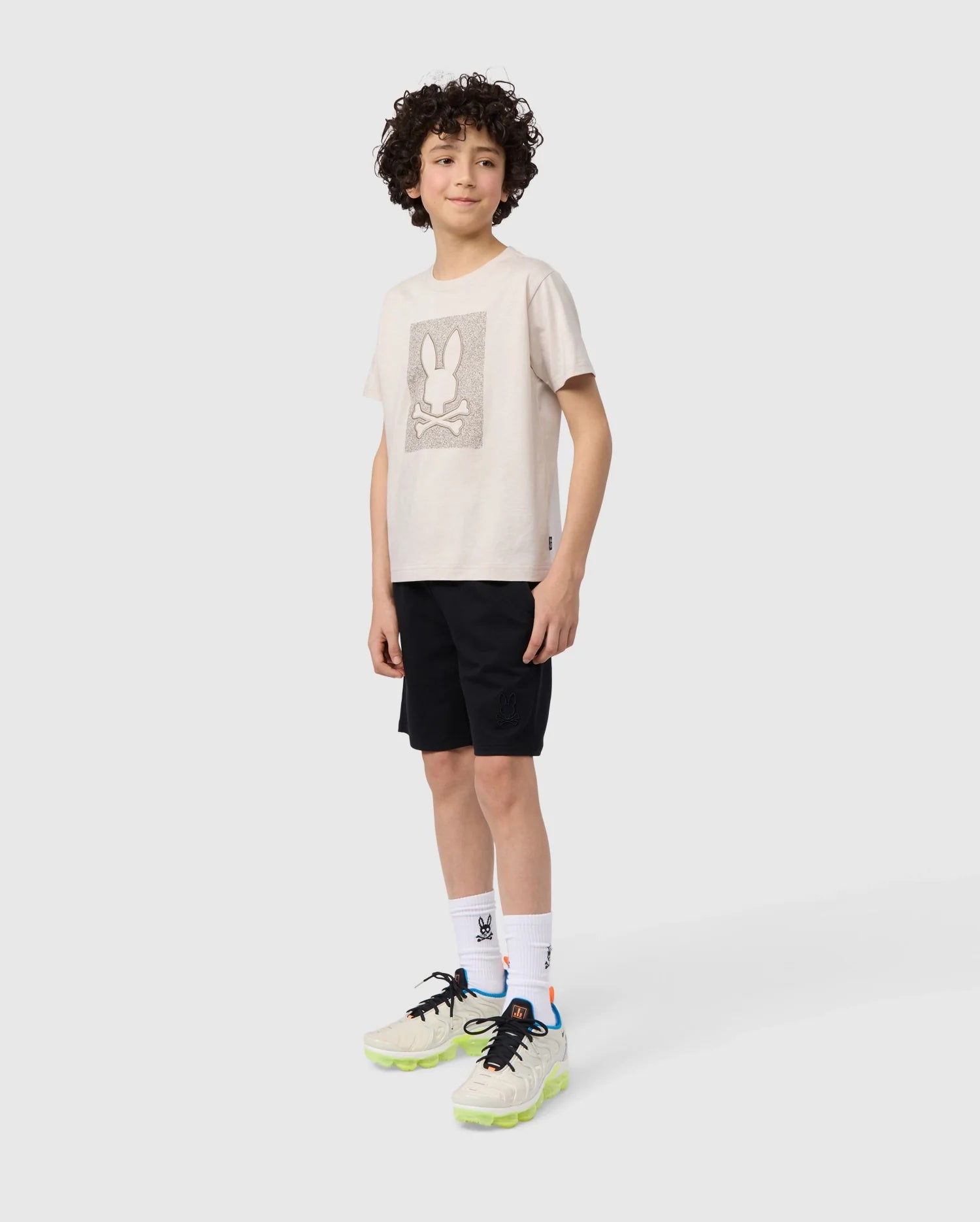 A young boy with curly hair stands against a plain white background. He is wearing a beige T-shirt with a bunny graphic, KIDS LIVINGSTON TERRY SWEATSHORT - B0R408B200 by Psycho Bunny for extra comfort, white socks with a blue logo, and beige sneakers with neon yellow soles. He is smiling and looking slightly off-camera.