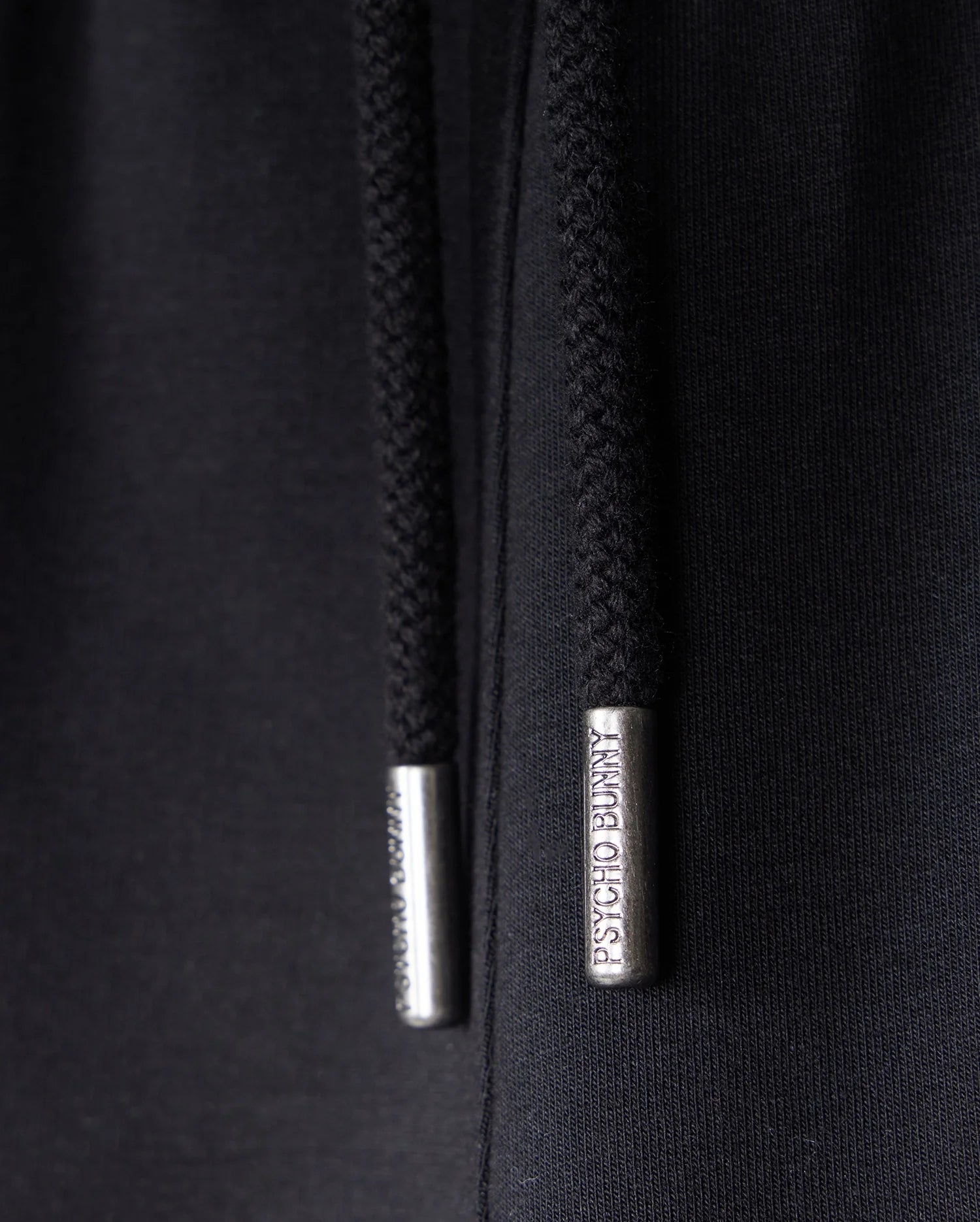 Close-up of black hoodie drawstrings with metallic aglets, crafted for ultimate comfort. The aglet on the right has "PSYCHO BUNNY" engraved on it, while the writing on the left aglet is partially obscured. Made from soft Micro French Terry material, it's perfect paired with KIDS LIVINGSTON TERRY SWEATSHORT - B0R408B200 by Psycho Bunny for a relaxed look.