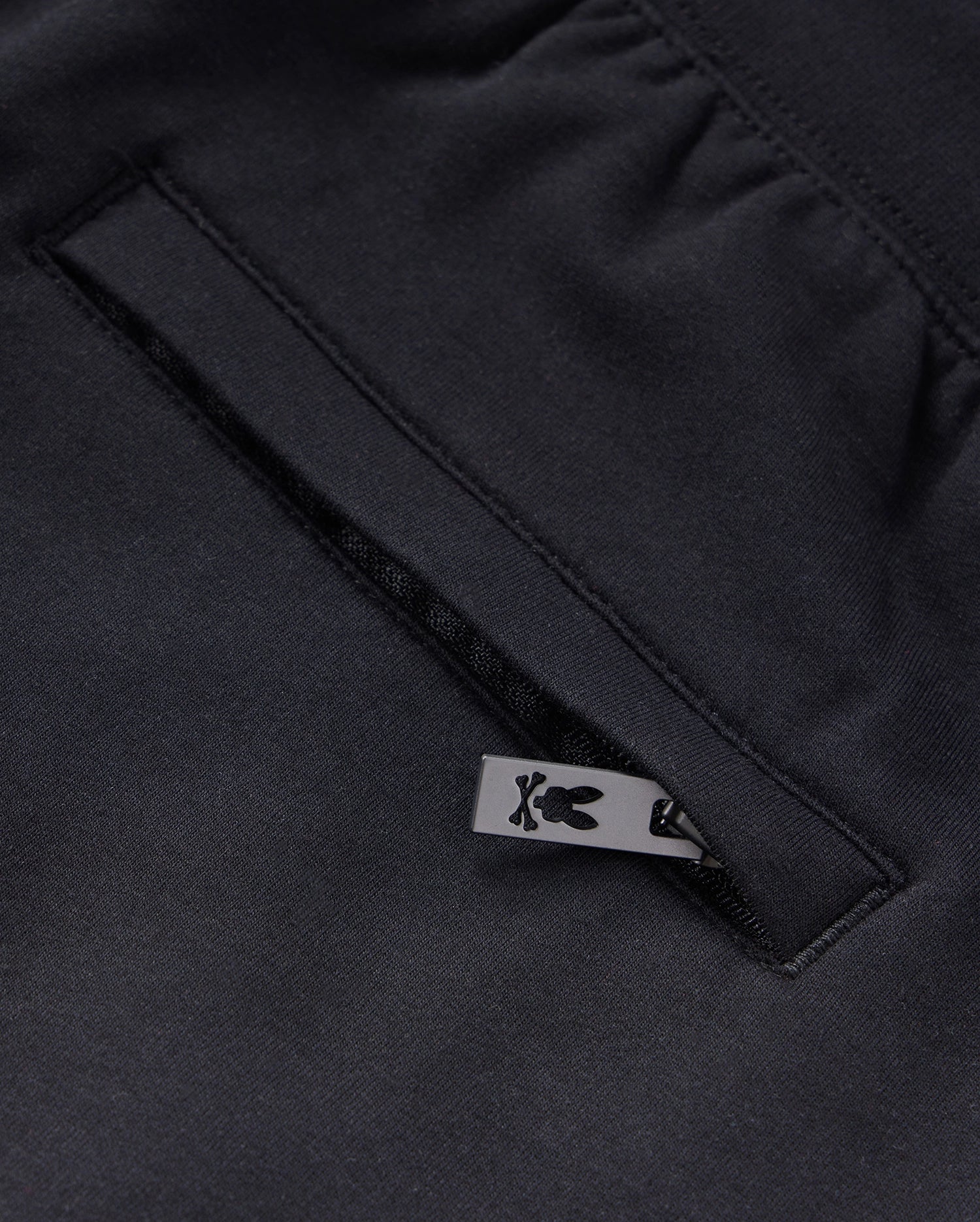 Close-up of a black KIDS LIVINGSTON TERRY SWEATSHORT - B0R408B200 by Psycho Bunny, showcasing a pocket with a neatly sewn opening. A small, black metal zip-pull with a bunny logo is partially visible, adding a touch of design detail to the garment. The fabric has a smooth and refined texture, enhancing comfort.