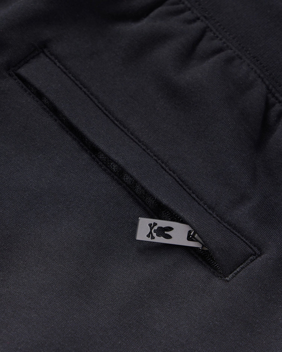 Close-up of a black KIDS LIVINGSTON TERRY SWEATSHORT - B0R408B200 by Psycho Bunny, showcasing a pocket with a neatly sewn opening. A small, black metal zip-pull with a bunny logo is partially visible, adding a touch of design detail to the garment. The fabric has a smooth and refined texture, enhancing comfort.