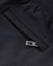 Close-up of a black KIDS LIVINGSTON TERRY SWEATSHORT - B0R408B200 by Psycho Bunny, showcasing a pocket with a neatly sewn opening. A small, black metal zip-pull with a bunny logo is partially visible, adding a touch of design detail to the garment. The fabric has a smooth and refined texture, enhancing comfort.